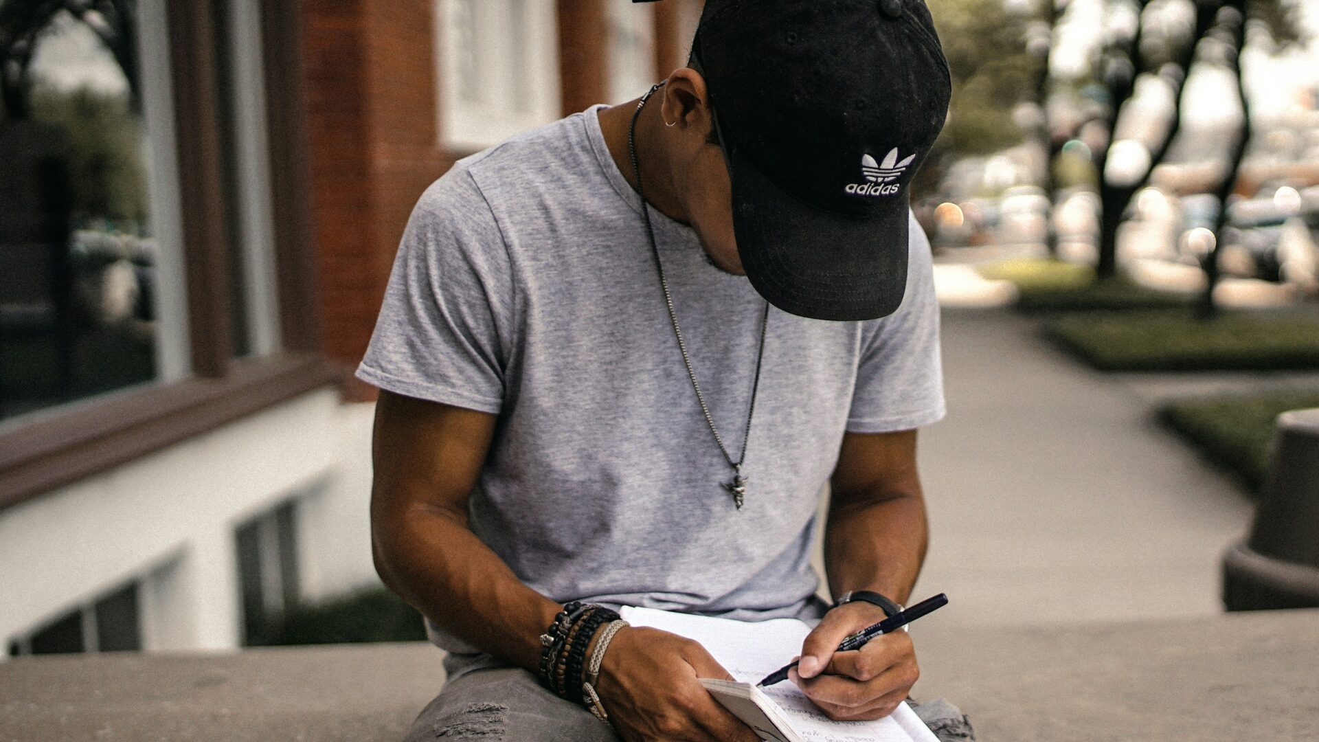 A young man wearing a black hat, sitting on a stone ledge and writing in his notebook.