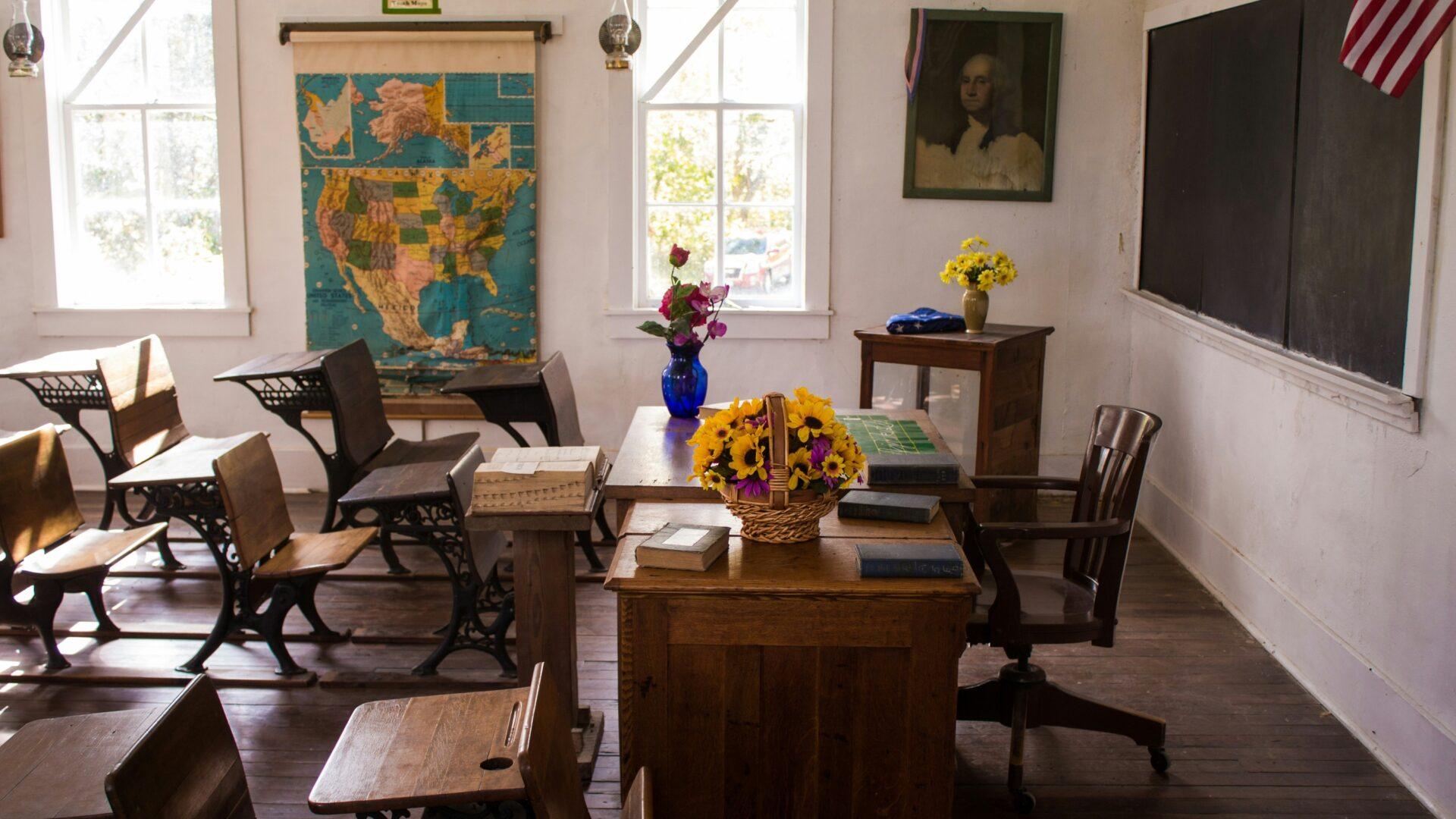 Elementary school classroom with a map on the wall, and the room filled with brown wooden desks and a teacher's desk with a bouquet of sunflowers on top, and bright sunlight coming through the window.