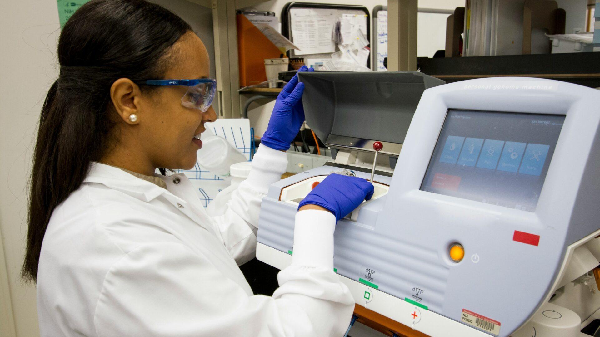 Young woman in a lab coat, wearing safety goggles and blue latex gloves working on a large medical machine in a lab setting